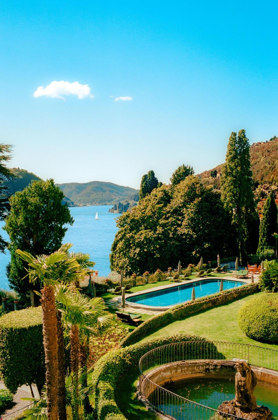 A luxurious garden with a swimming pool and fountain overlooks a large lake with mountains in the distance.