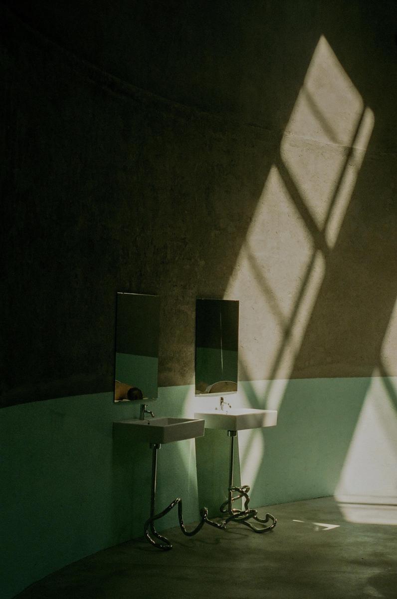Two sinks with mirrors are illuminated by a geometric pattern of sunlight on a wall.