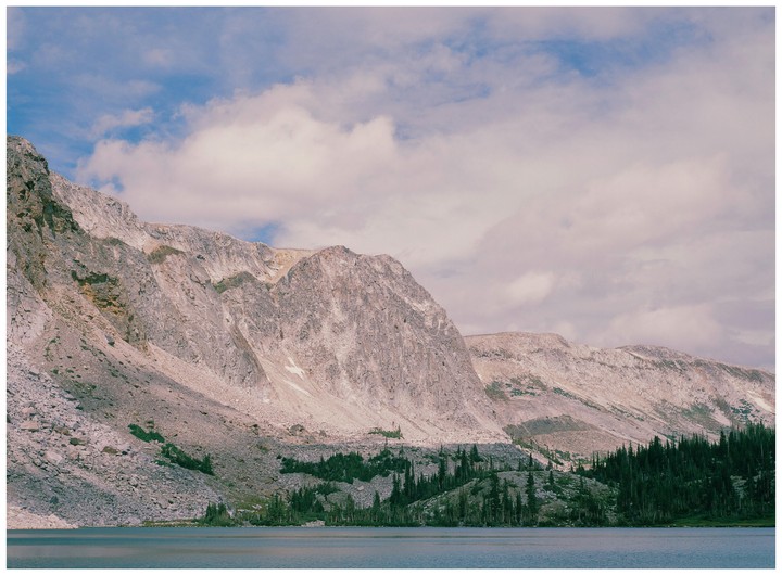 A mountain range with sparse trees rises from the edge of a lake under a cloudy sky.
