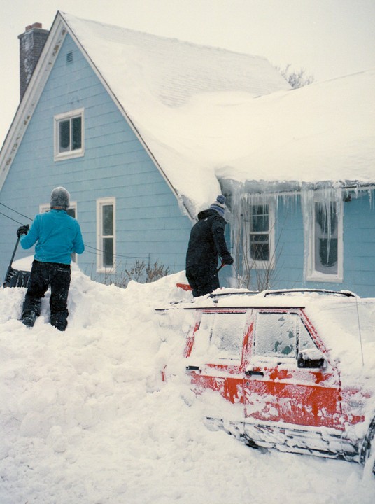 Two people shovel snow from around a red car and a blue house with icicles hanging from the roof.