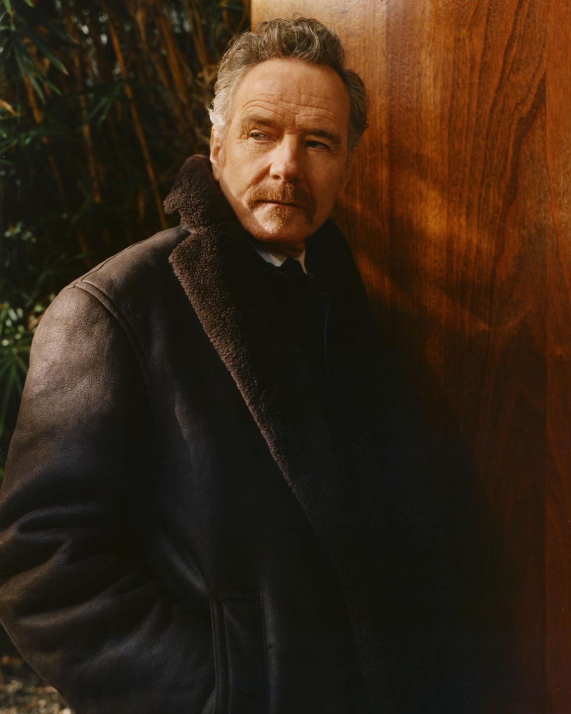 A man with a mustache wears a dark coat with a fur collar and leans against a wooden surface.