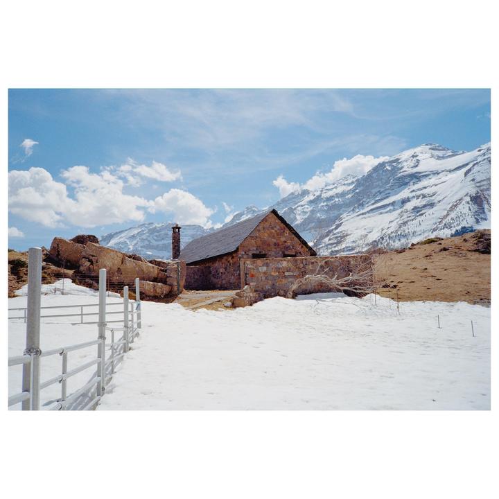 A stone building with a dark roof and chimney stands in a snowy landscape with mountains in the background.