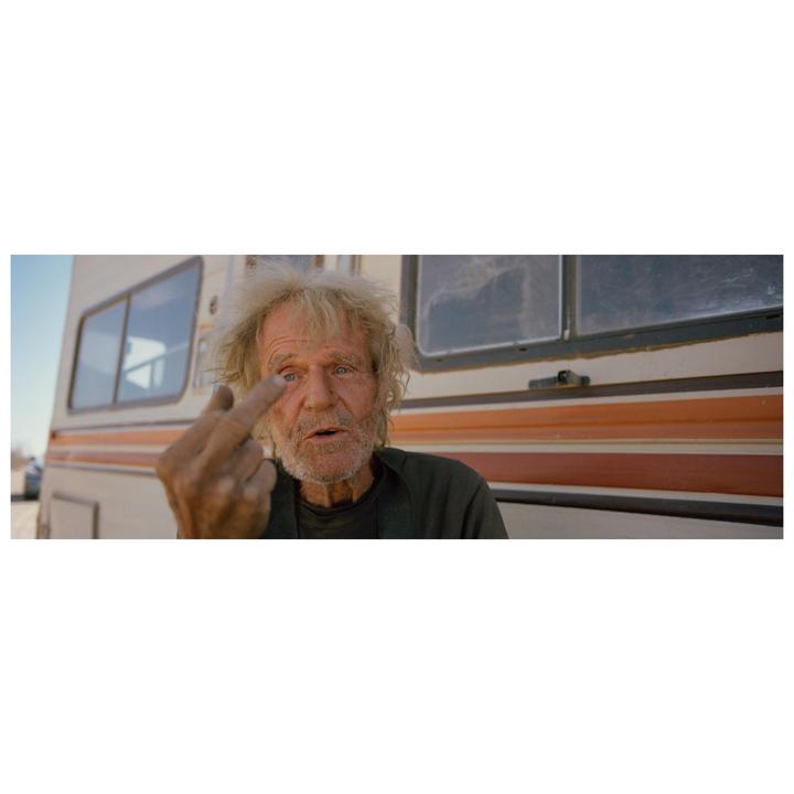 A man with a beard and messy hair makes an obscene gesture in front of a recreational vehicle.