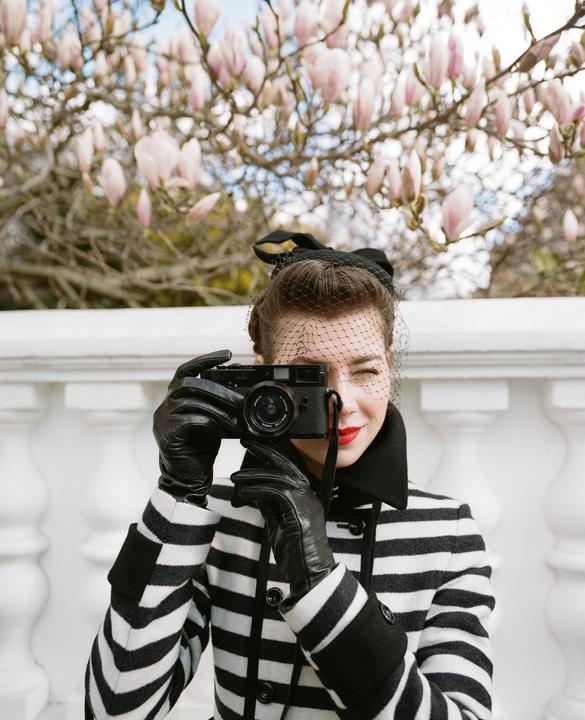 A woman in a striped coat and veil hat holds a camera to her eye, with magnolia blossoms in the background.