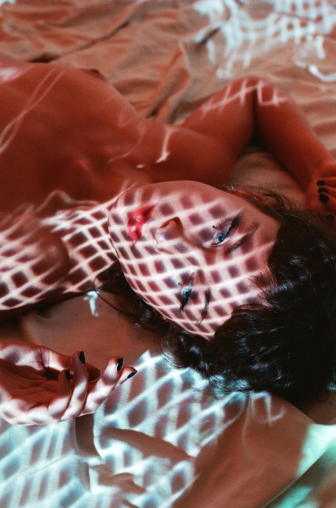 A woman with dark hair and red lipstick lies on a bed with a diamond pattern of light and shadow cast across her body.