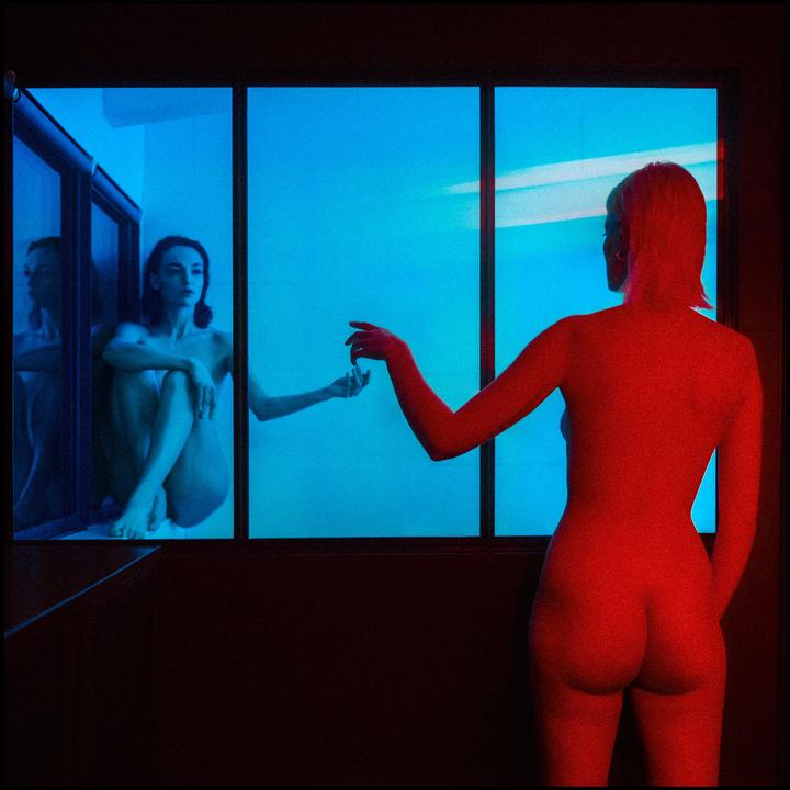 A nude woman with red skin stands with her back to the viewer, reaching towards a nude woman with blue skin sitting in a window.
