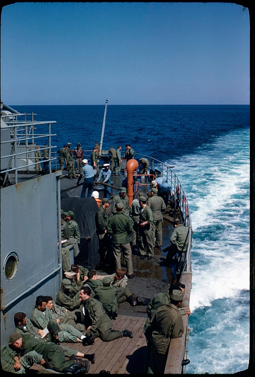 A group of men in military uniform are gathered on the deck of a ship, with the ocean visible in the background.