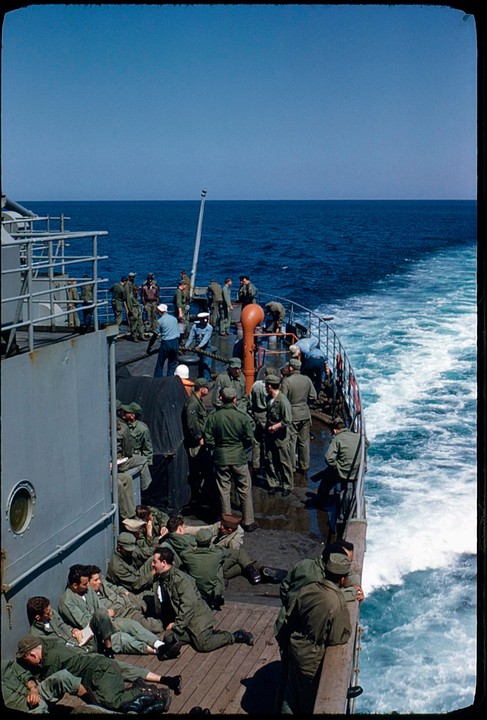 A group of men in military uniform are gathered on the deck of a ship, with the ocean visible in the background.