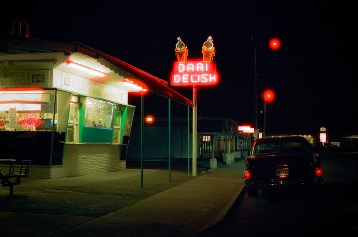 A neon sign for Dari Delish ice cream stands next to a brightly lit ice cream stand and a car at night.