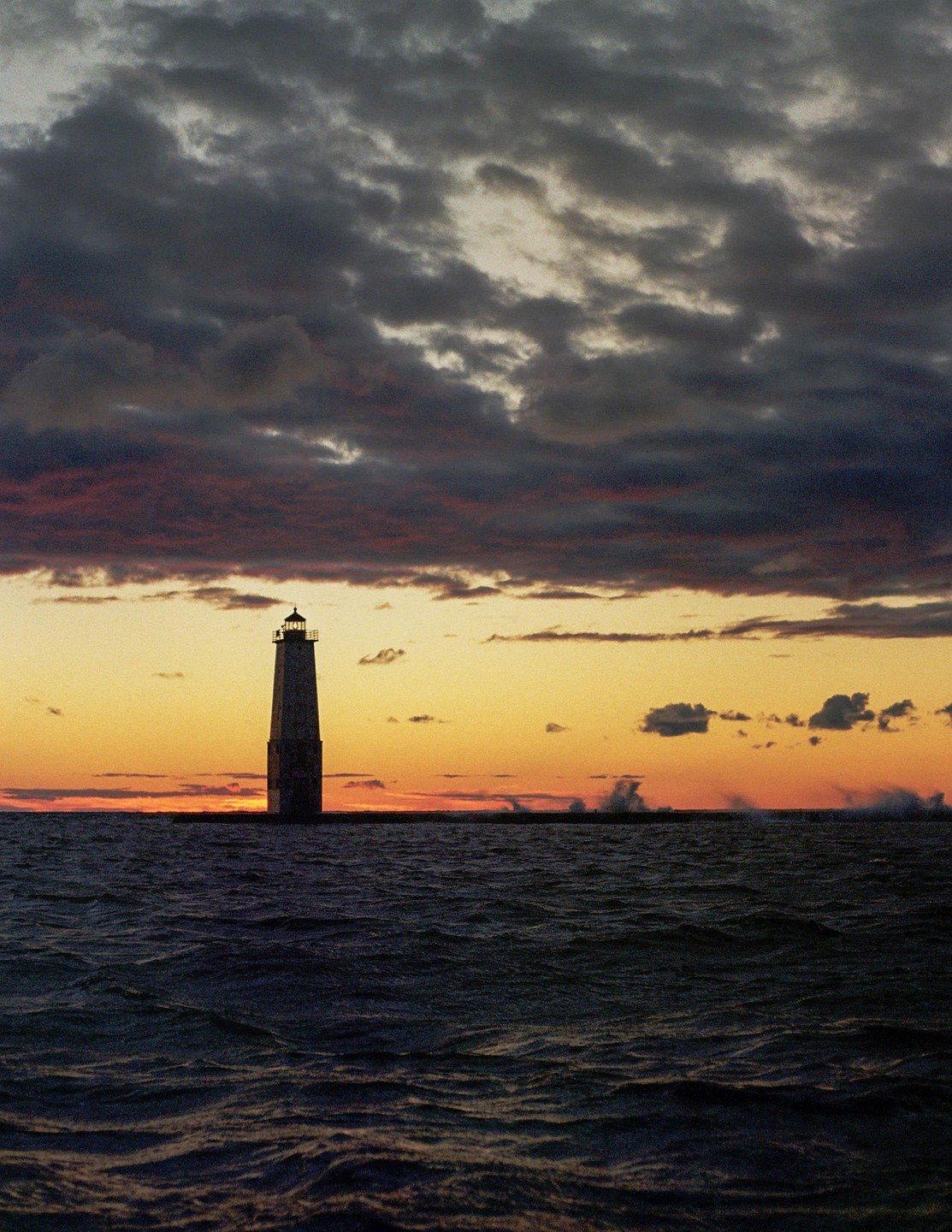 A lighthouse stands in the water under a dramatic sky at sunset.
