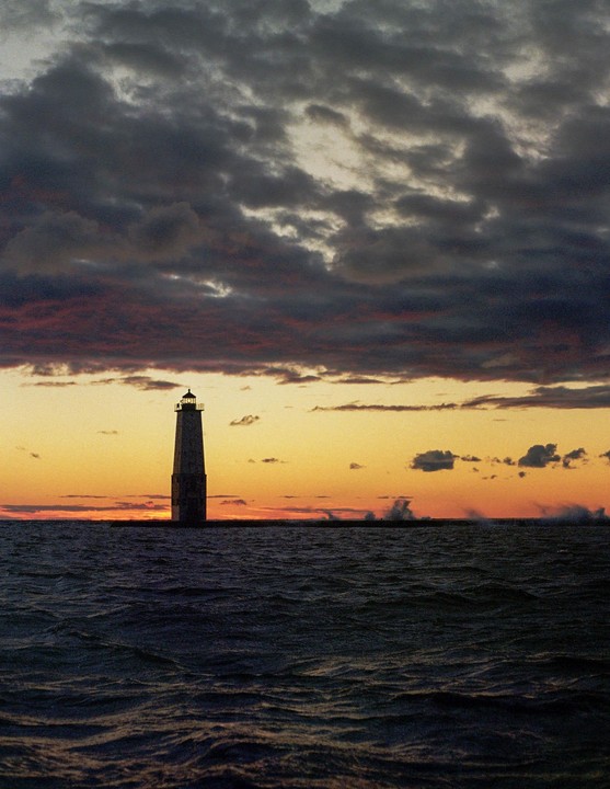 A lighthouse stands in the water under a dramatic sky at sunset.