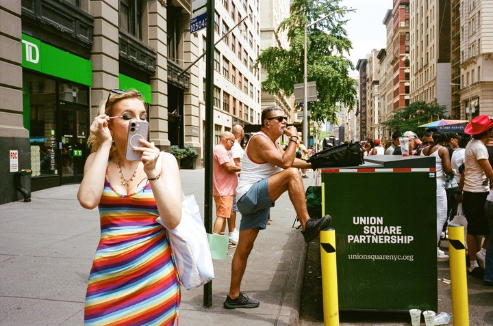 A woman in a rainbow dress applies makeup using her phone as a mirror on a city street with other people and buildings in the background.