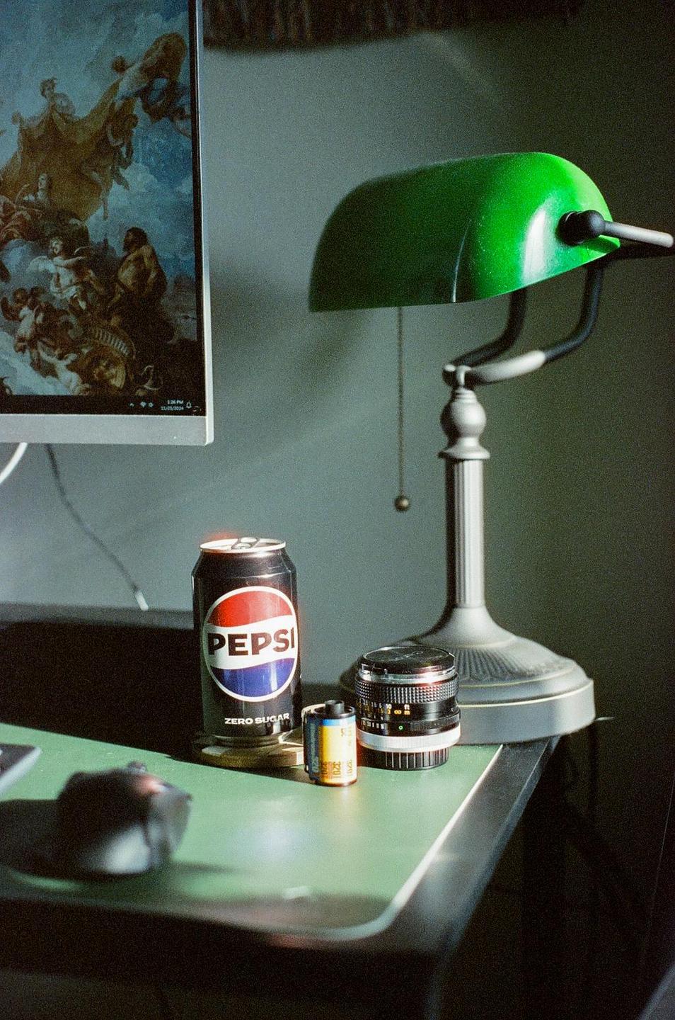 A Pepsi can, a roll of film, and a camera lens sit on a green desk next to a green lamp and a computer monitor displaying a painting.