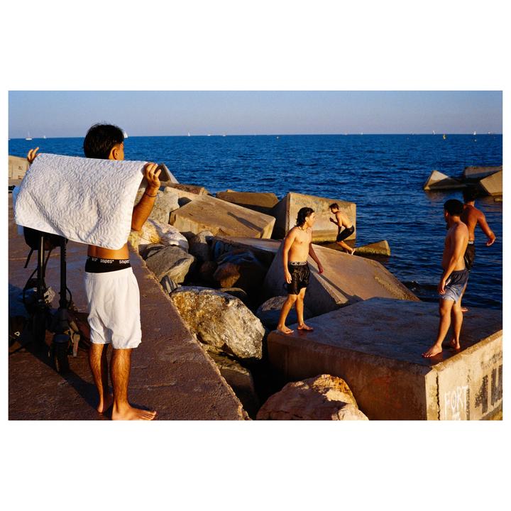 A group of young men are gathered on concrete blocks by the sea, with one holding a white towel over his head.