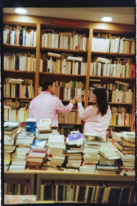 A couple stands in a bookstore aisle, touching hands over a shelf of books.