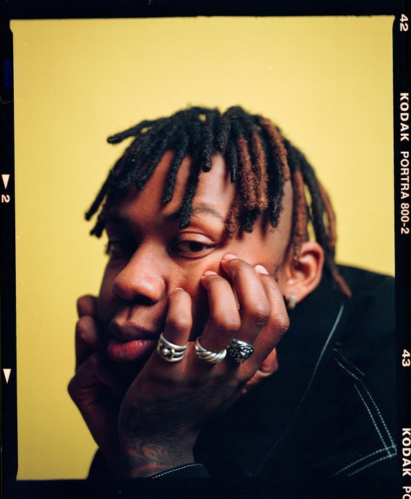 A man with dreadlocks rests his head on his hand, wearing rings on his fingers.