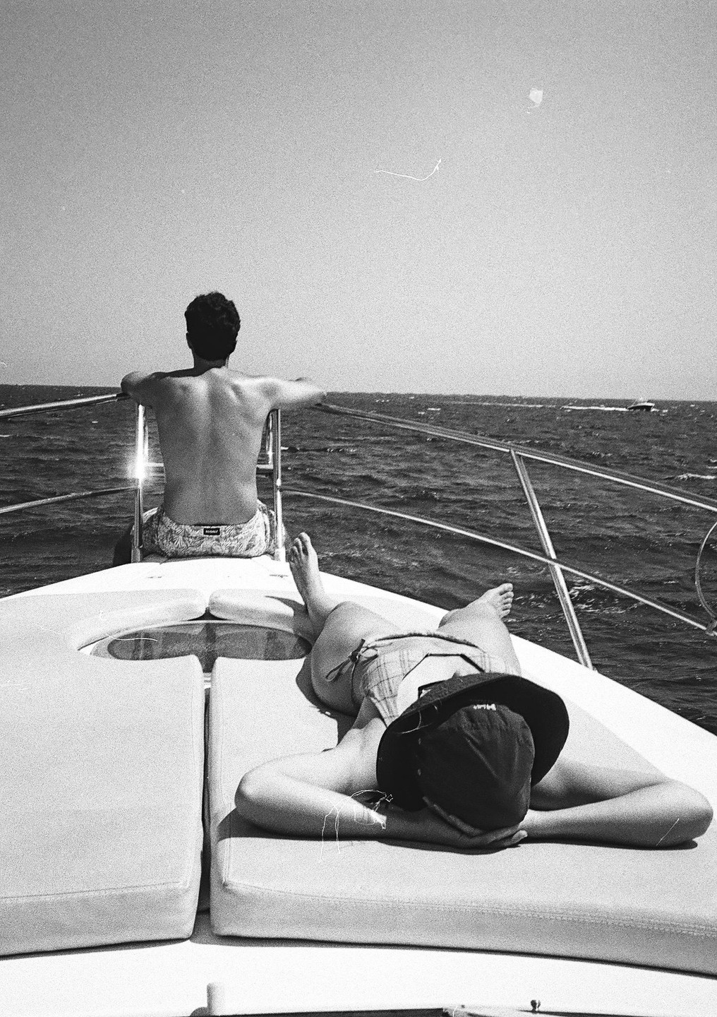 A man sits at the bow of a boat looking out at the ocean while a woman sunbathes in front of him.