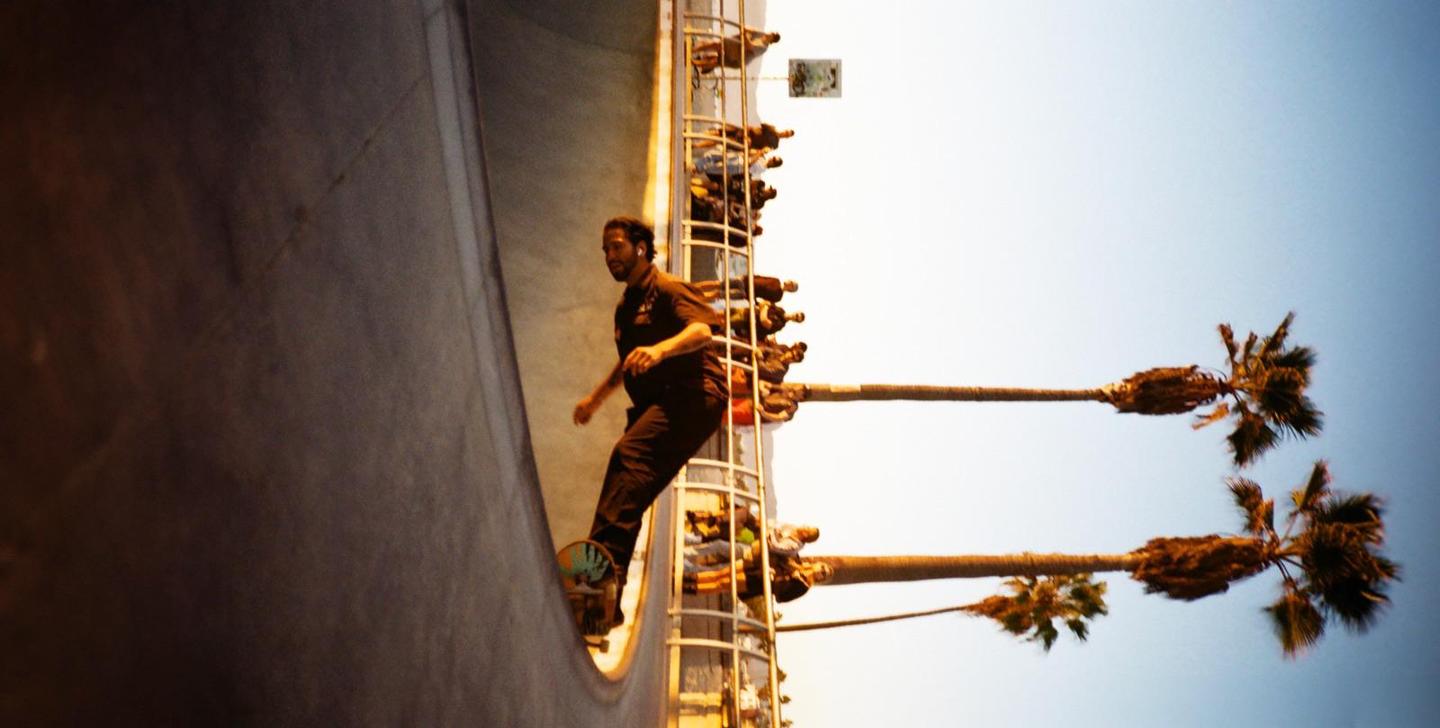 A man on a skateboard is in a skate park with palm trees and spectators in the background.