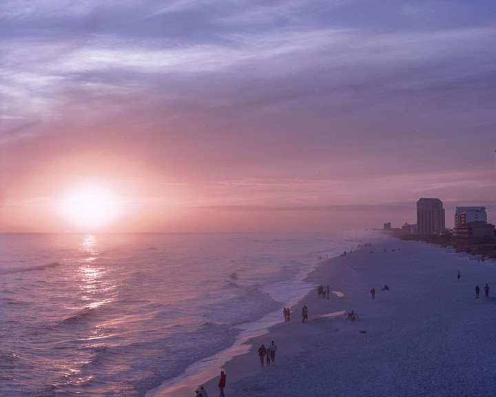 The sun sets over a beach with people walking along the shore and buildings in the distance.