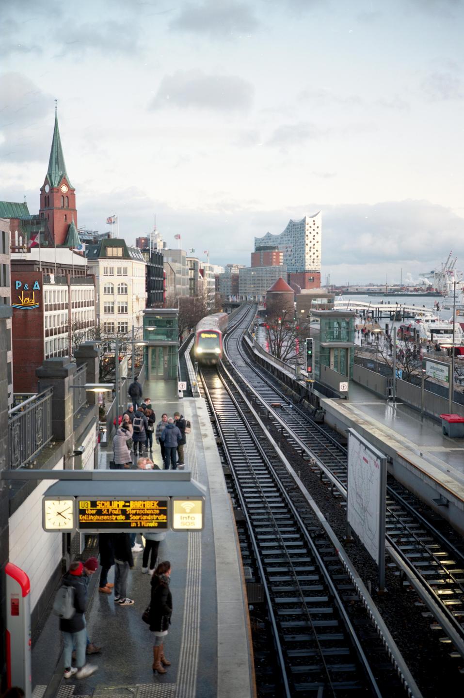 A train approaches a station platform where people are waiting, with a city skyline and harbor in the background.