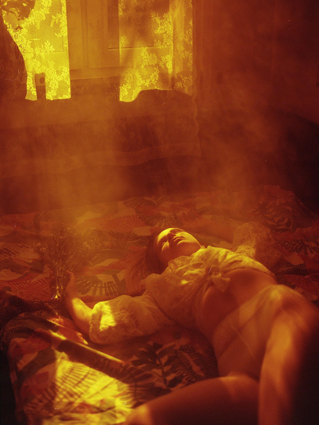 A woman lies on a patterned bedspread, holding a small bouquet of flowers, with golden light streaming through a window.