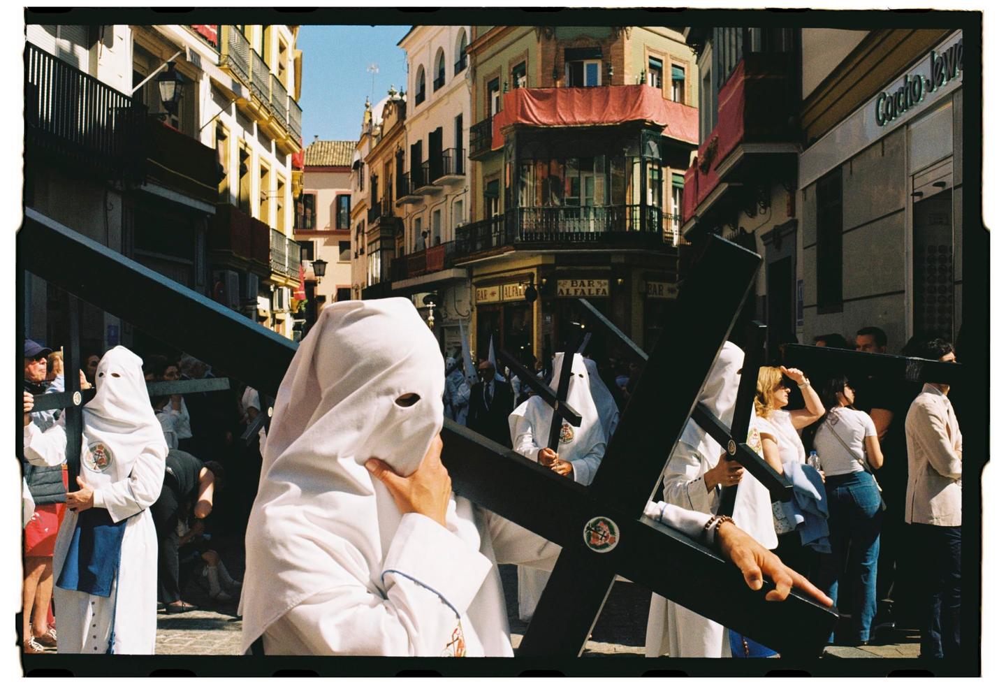 People in white robes with pointed hoods carry large black crosses down a sunny street lined with buildings.
