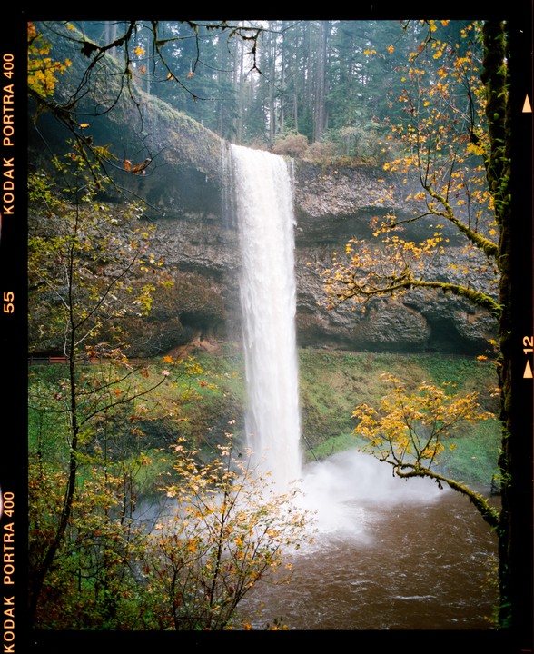 A tall waterfall cascades into a pool surrounded by trees with autumn foliage.