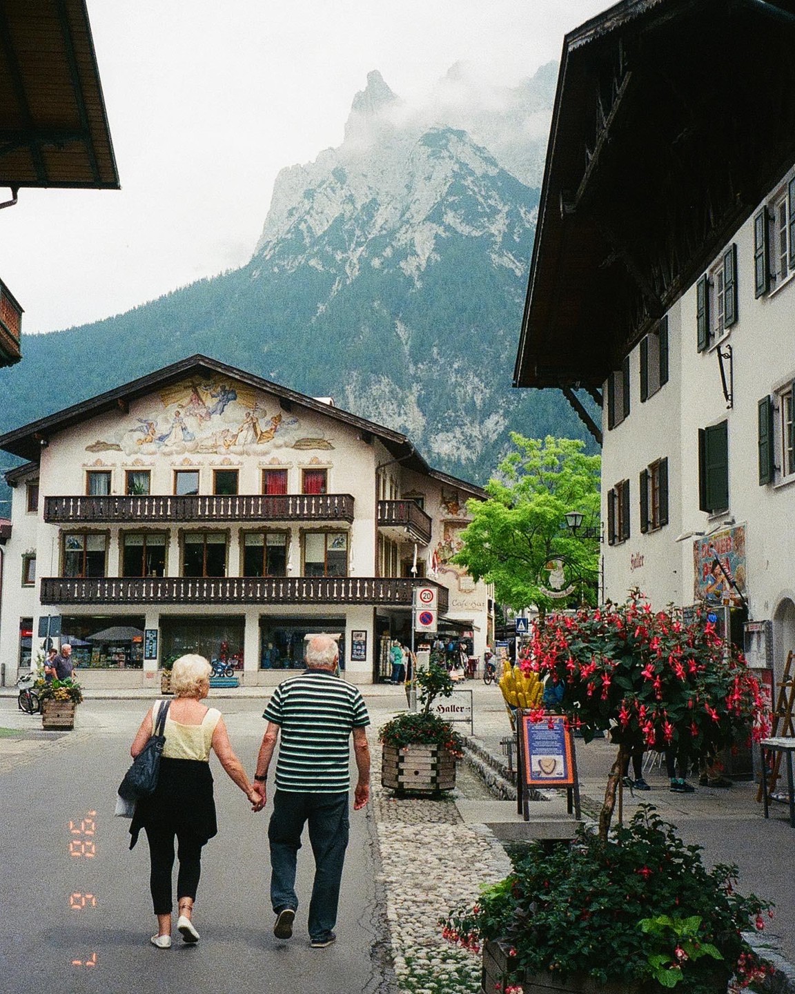 A couple walks hand-in-hand down a street in a mountain town with buildings featuring painted facades and balconies.