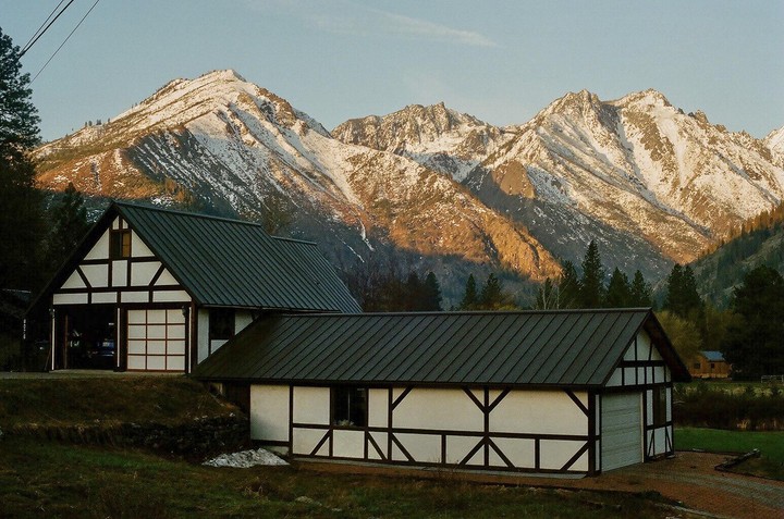 Two white buildings with dark roofs and exposed timber frames sit in front of snow-capped mountains.