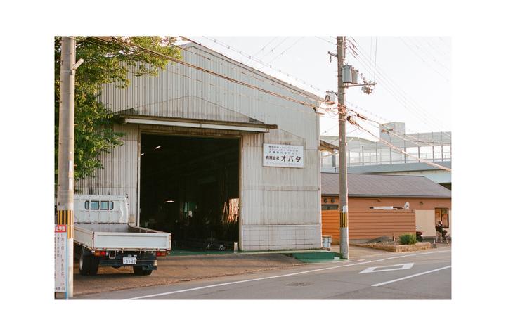 A white flatbed truck is parked in front of a large industrial building with its garage door open, next to a street with power lines overhead.