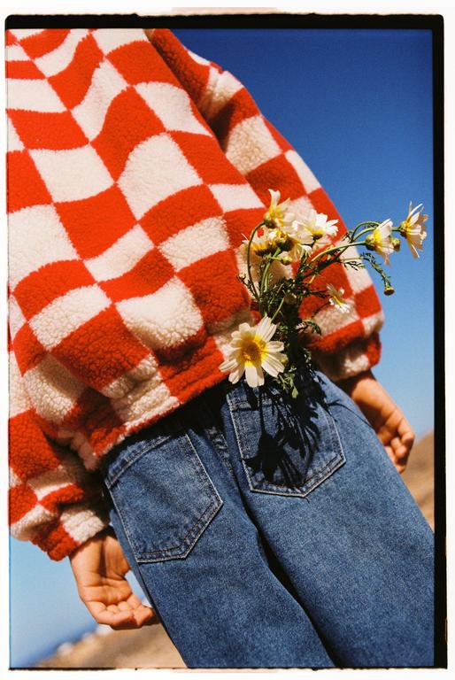 A person wearing a red and white checkered jacket and blue jeans has white flowers tucked into a back pocket.