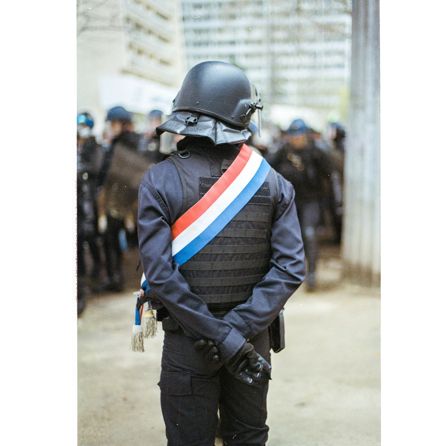 A police officer wearing a helmet and a sash with the colors of the French flag stands with hands clasped behind their back.