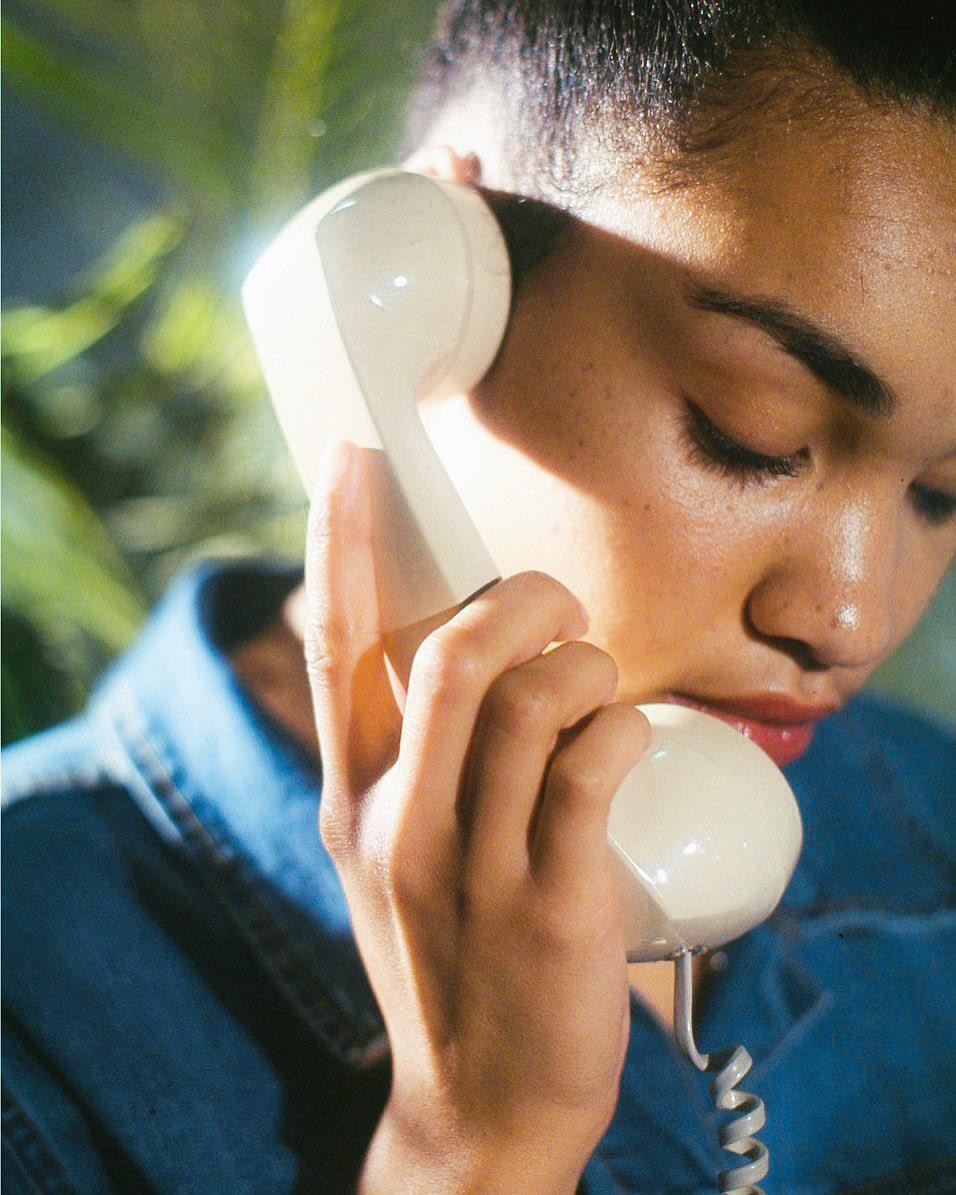 A woman holds a white telephone receiver to her ear, with her eyes closed and a shadow across her face.