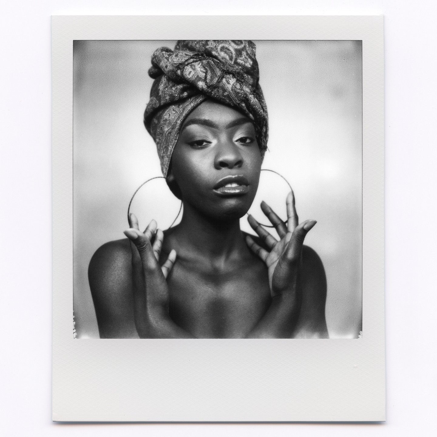 A woman with a patterned head wrap and large hoop earrings poses with her hands raised.