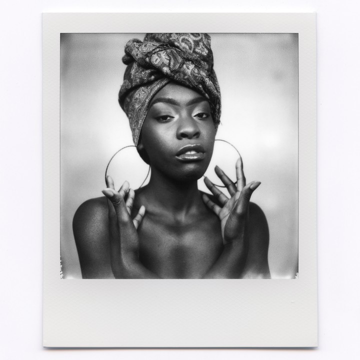 A woman with a patterned head wrap and large hoop earrings poses with her hands raised.