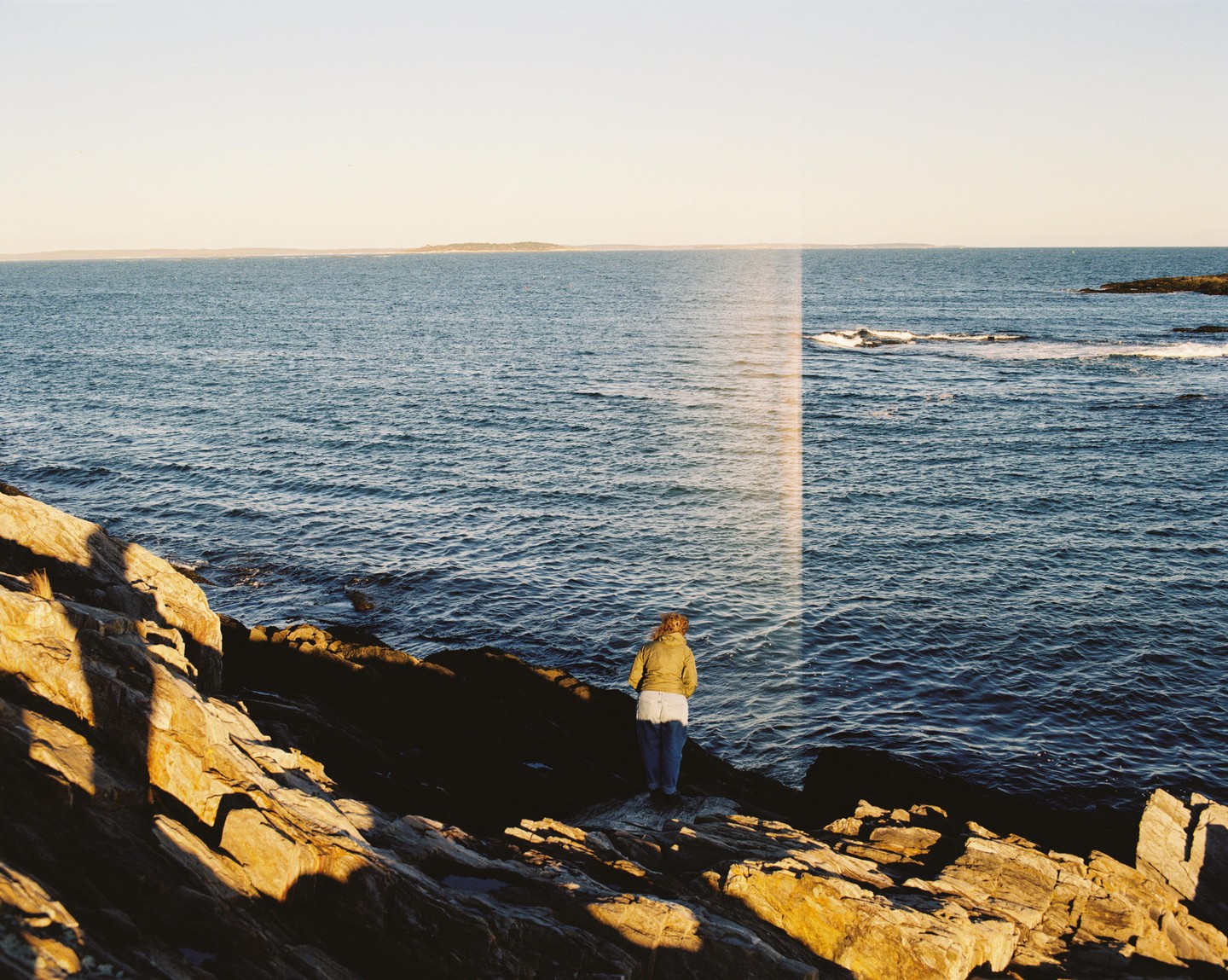 A woman stands on a rocky shore looking out at the ocean under a clear sky.
