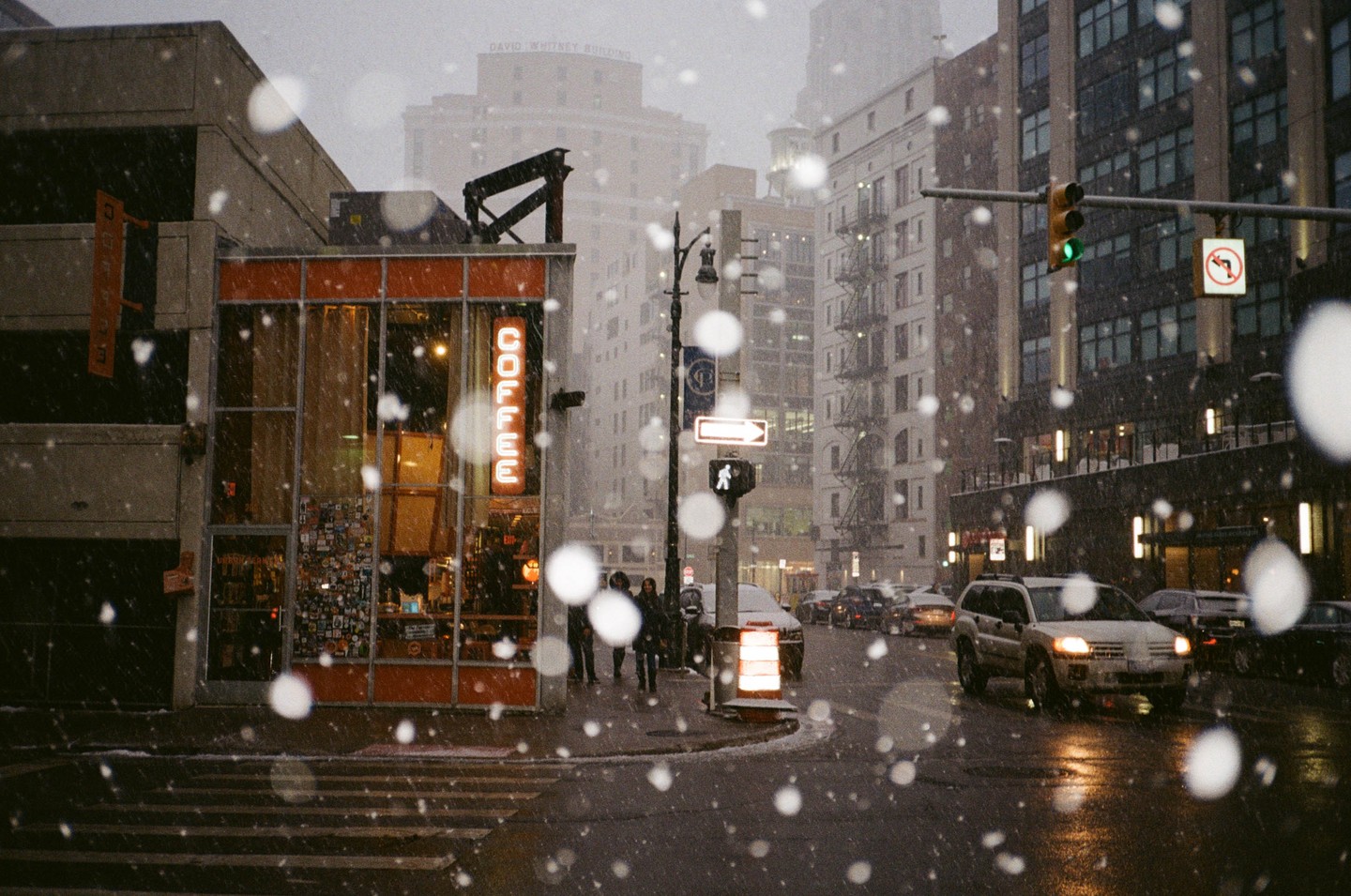 A street corner with a coffee shop and cars driving by during a snowfall in a city.
