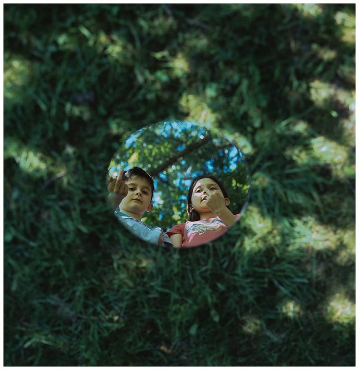 Two children are reflected in a circular mirror placed on a grassy surface.
