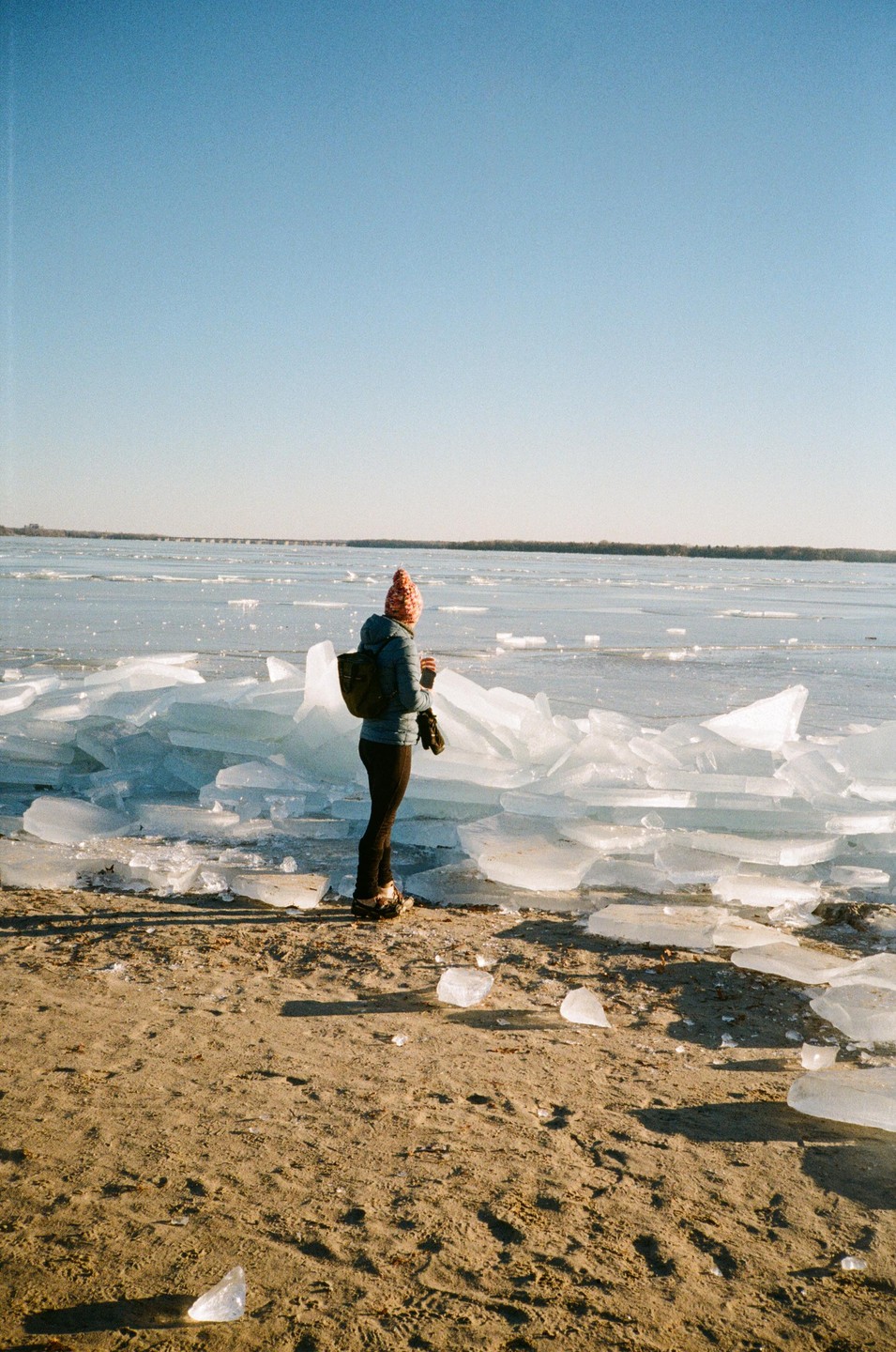 A woman stands on a sandy beach next to a frozen lake with large chunks of ice.