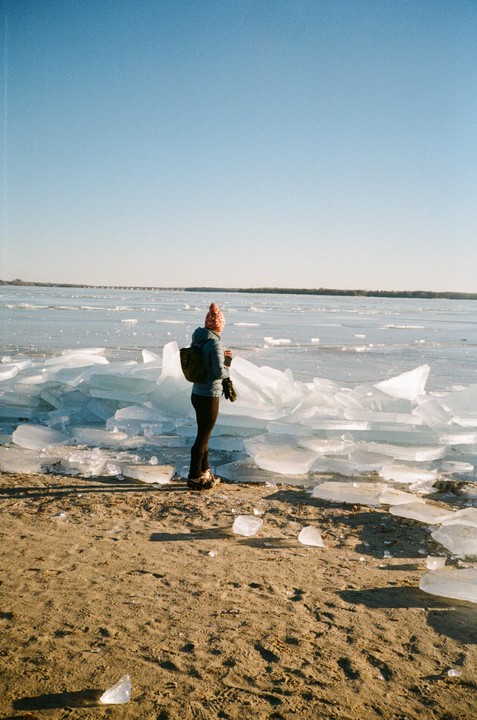 A woman stands on a sandy beach next to a frozen lake with large chunks of ice.