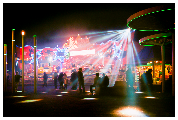 People gather at night around a brightly lit fairground ride with star shapes and neon lights.