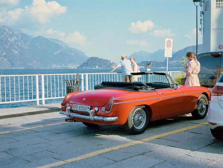 A red convertible car is parked by a lake with mountains in the background and people standing near a railing.