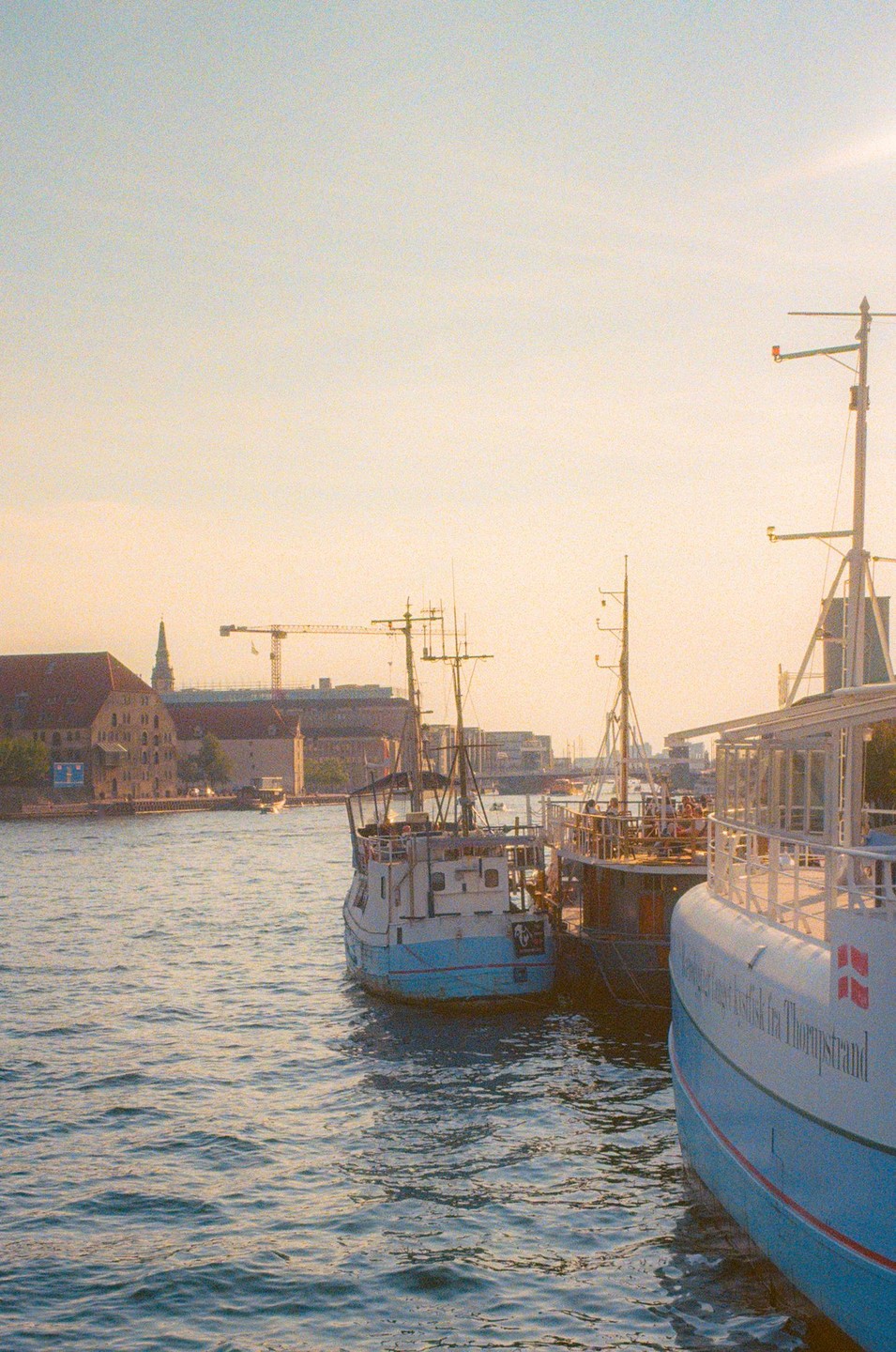 Three boats are moored in a harbor with buildings and a crane visible in the background.