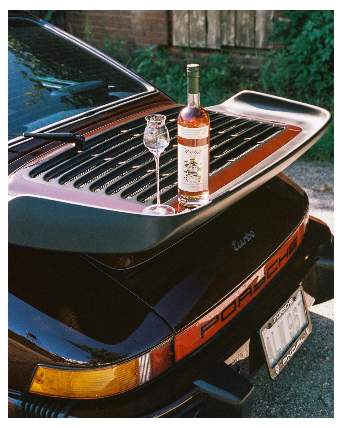 A bottle of liquor and a snifter glass sit on the spoiler of a black Porsche.
