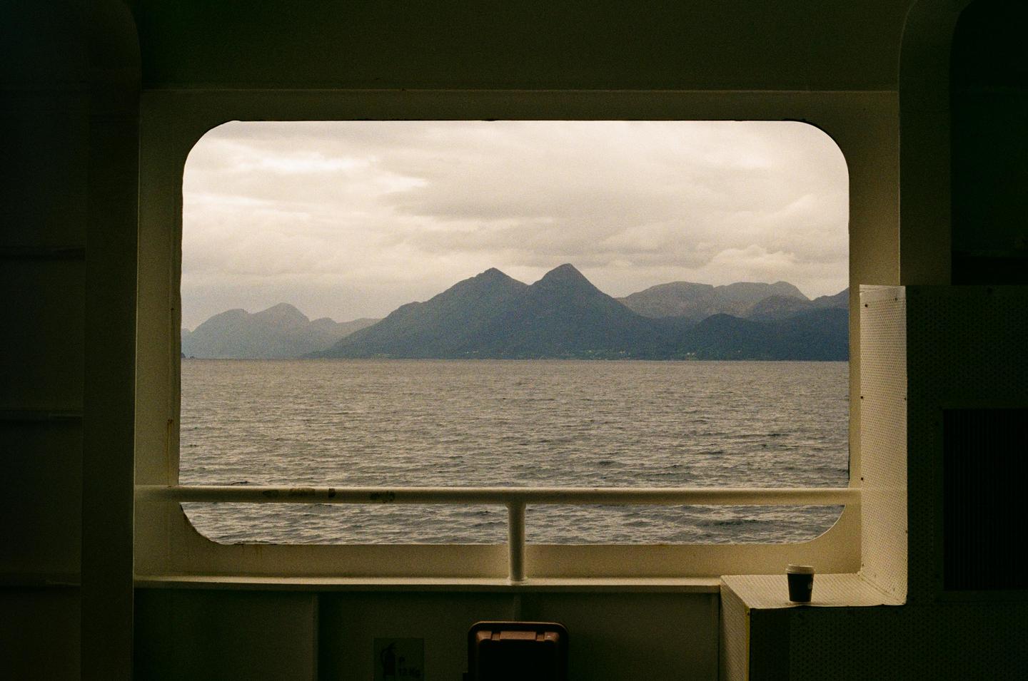 A view of mountains and water from a window on a boat, with a cup on a ledge in the foreground.