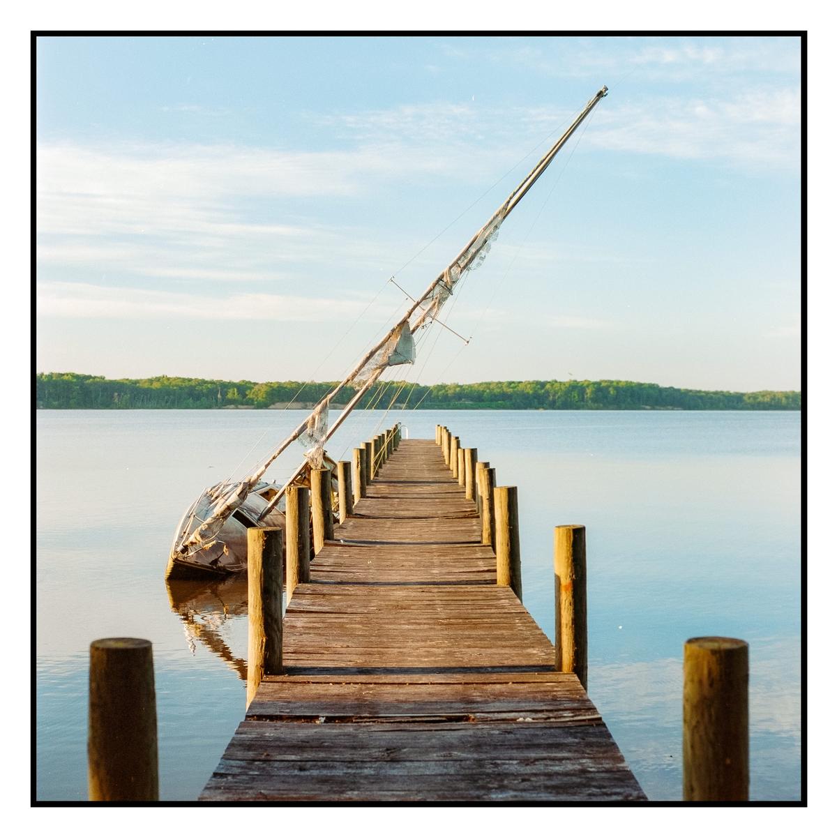 A wooden pier extends into a body of water where a partially submerged sailboat wreck is visible.