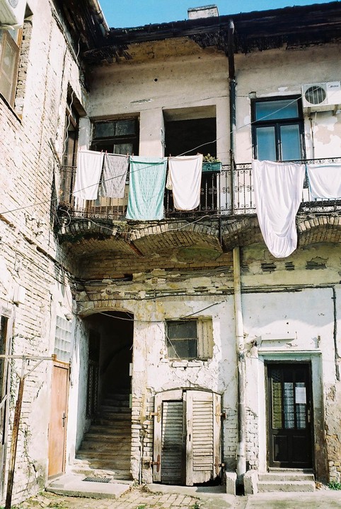 Laundry hangs on a balcony above a courtyard with crumbling walls and multiple doorways.
