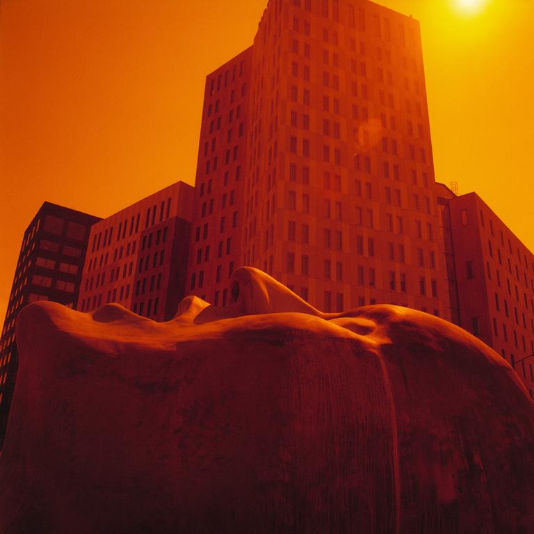 A large sculpture of a face lies in front of a tall building under an orange sky.
