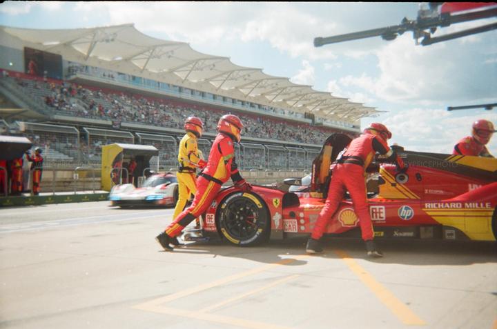 Three men in racing suits push a red race car on a track with a grandstand full of spectators in the background.