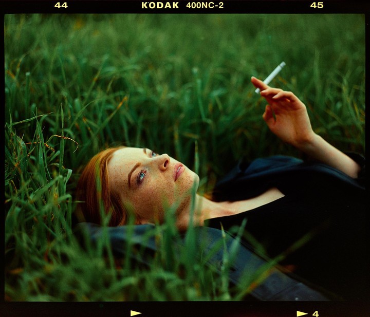 A woman with red hair and freckles lies in tall green grass, holding a cigarette.
